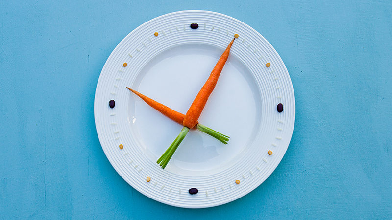 Clock made with two carrots as hands on a white plate, with rice and seeds for numbers.