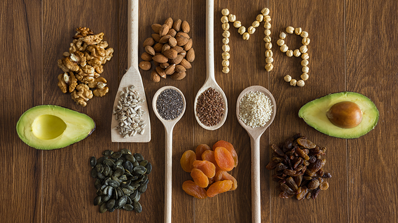 Aerial view of various nuts, seeds and fruits on a dark brown, wooden table with Mg spelled out in nuts. 