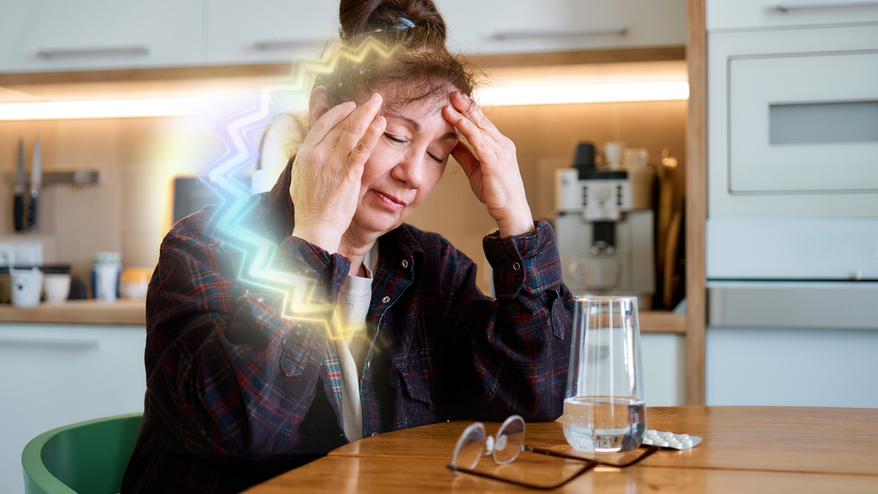 A person with brown hair and their eyes closed sits at a kitchen table with hands on their head, representing an ocular migraine. There is a colorful zigzagging pattern in the image, often seen with an ocular migraine.