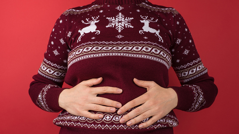 Person's torso wearing a maroon sweater with white reindeer and snowflake patterns on it. The person is holding their stomach as if full..