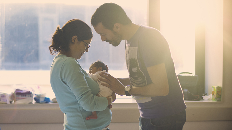 Parents holding a newborn with the sun streaming in the window behind them.