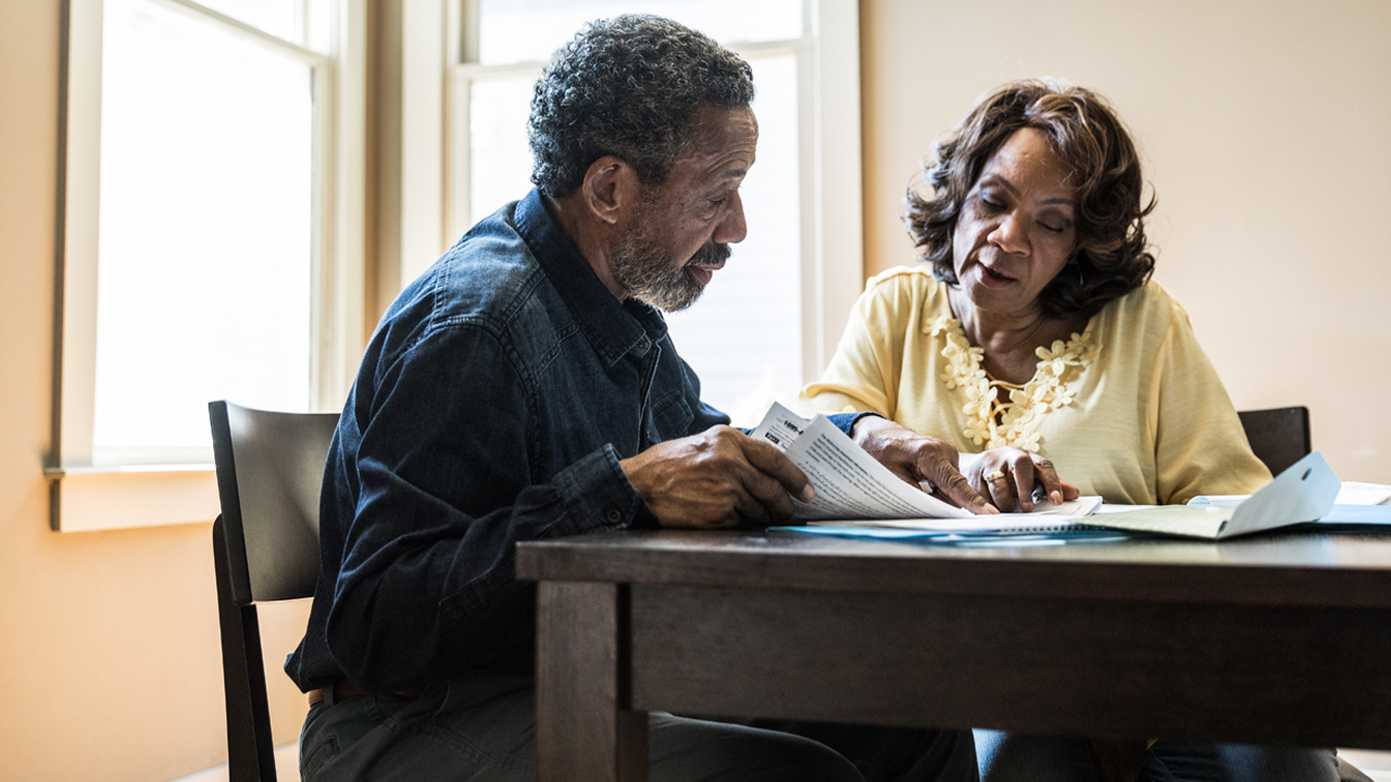 Two mature people at a kitchen table looking at and discussing documents.