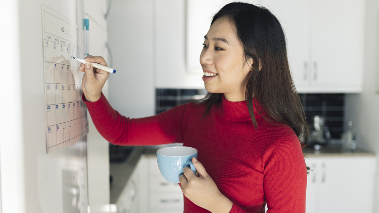 Smiling person with long black hair and red turtleneck circles a date on a whiteboard calendar and holds a cup of coffee.