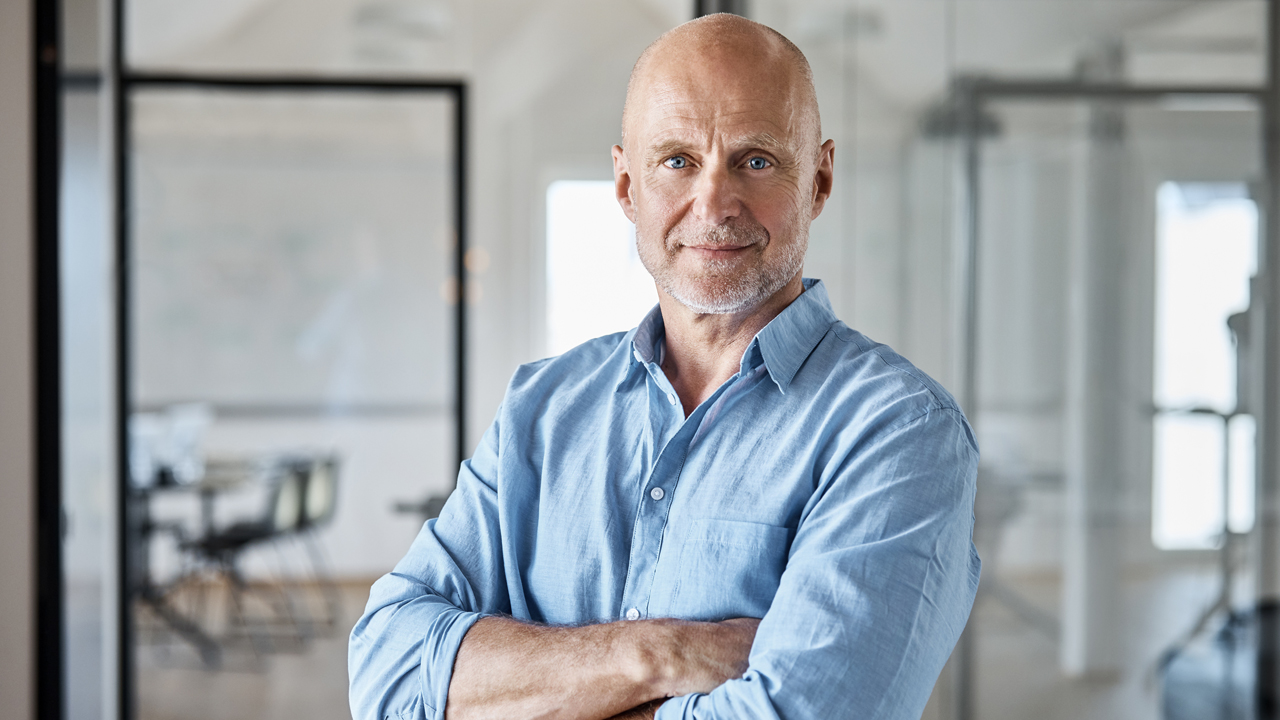 Man in blue Oxford shirt standing in modern office with crossed arms.