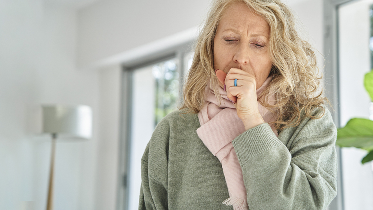 An older adult wearing a green sweater and a pink scarf putting hand on mouth while coughing.