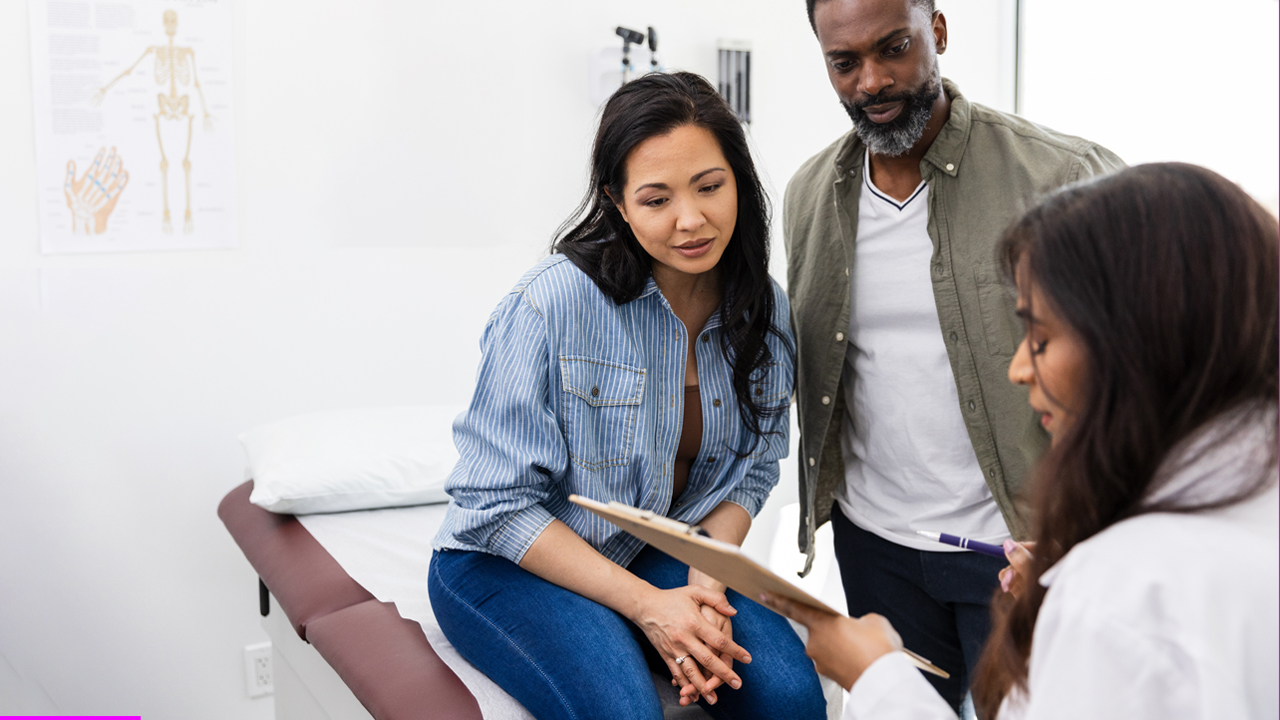 Person with long black hair sits down next to a person with short black hair who is standing in a doctor’s office. Another person with long black hair wearing a white doctor’s coat holds up a clipboard.