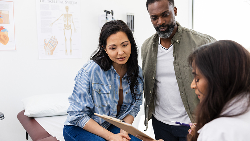 Person with long black hair sits down next to a person with short black hair who is standing in a doctor’s office. Another person with long black hair wearing a white doctor’s coat holds up a clipboard.