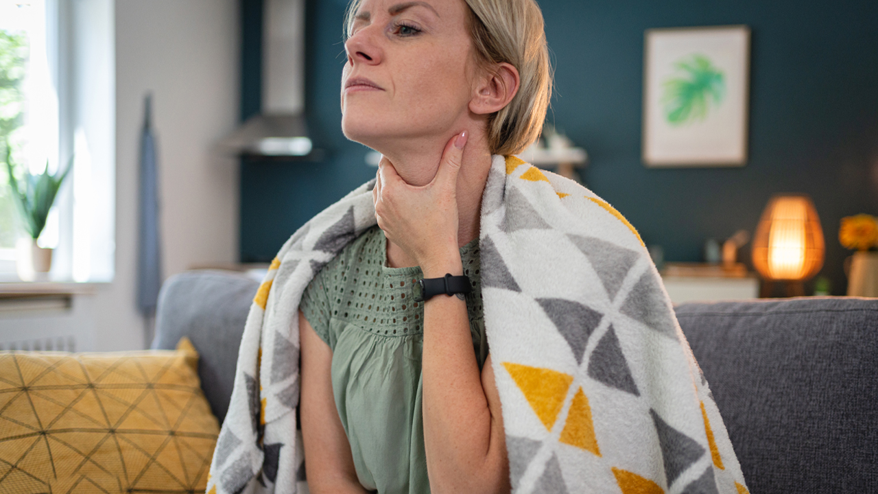 Person touching their throat while sitting on a couch, wrapped in a colorful blanket.
