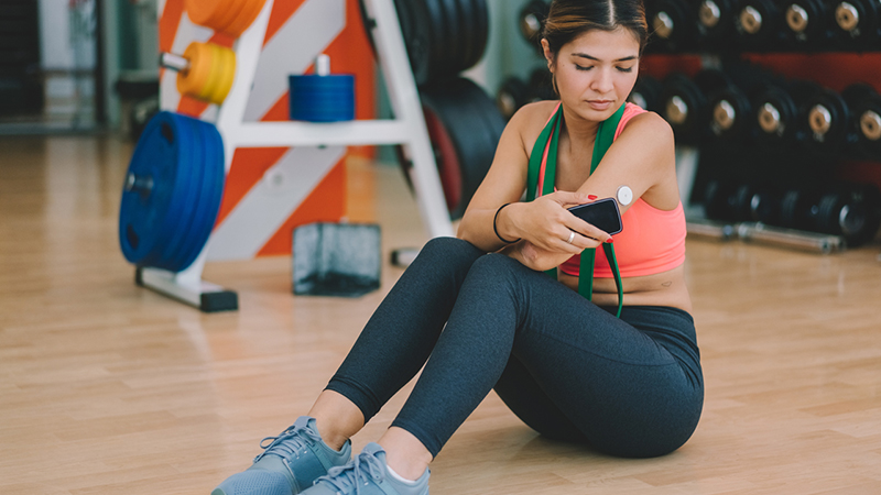 A woman in workout clothes sits on the floor in a gym and measures her blood sugar levels with a phone and diabetes sensor on her arm.