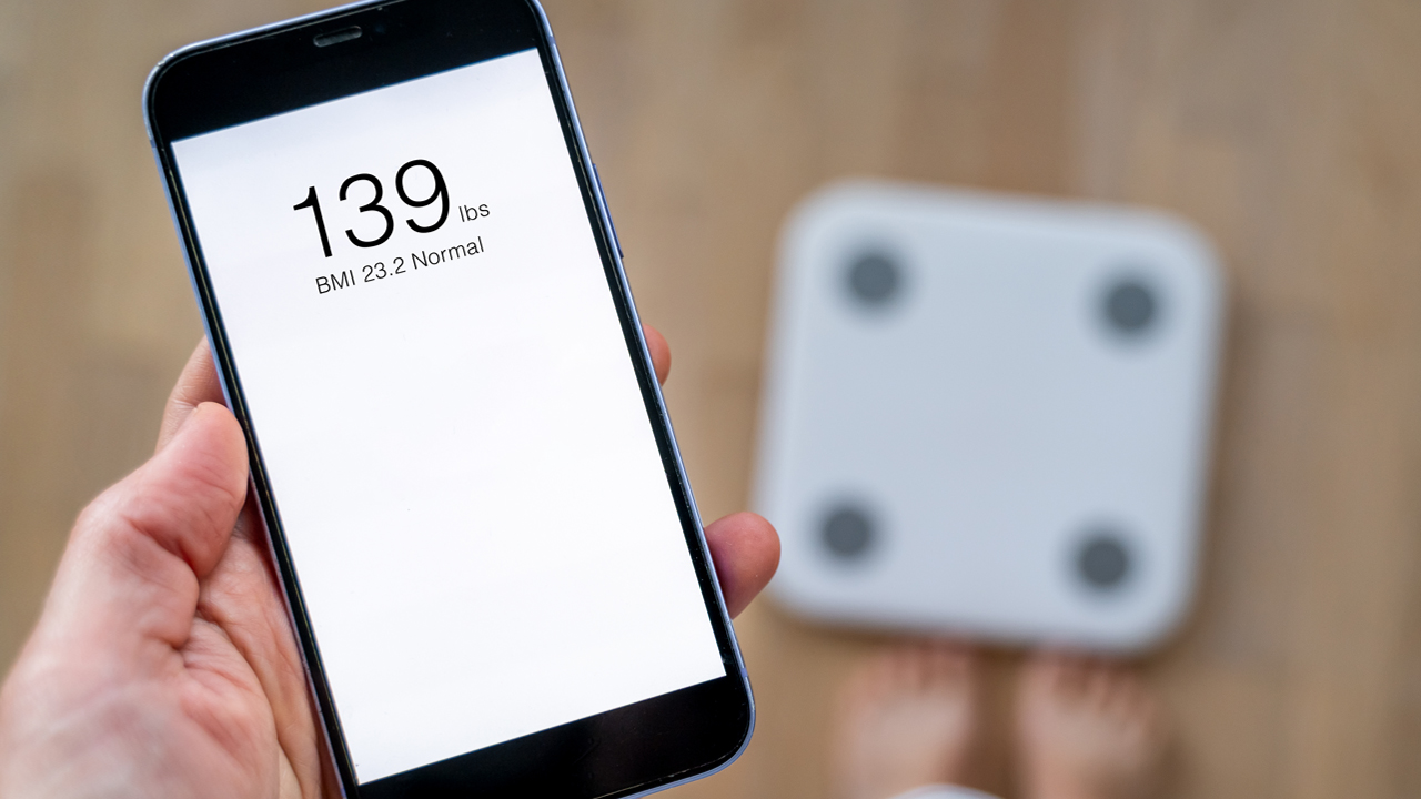 Person standing in front of a scale and holding a smart phone showing their weight and body mass index 