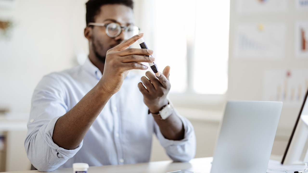 A man takes a blood sample from his index finger tip with a lancet pen to test blood sugar levels, sitting at a desk in an office with a laptop in the foreground.