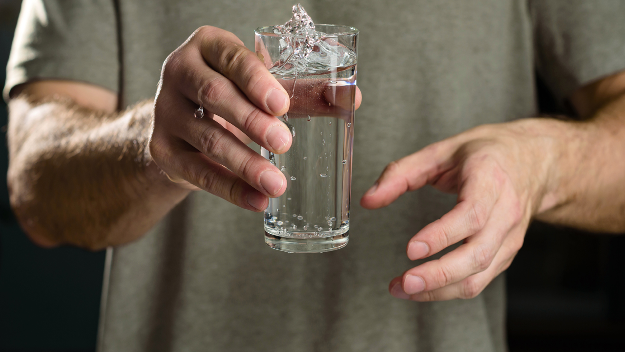Person pictured from the shoulders down holding a glass of water that looks like it is about to spill.