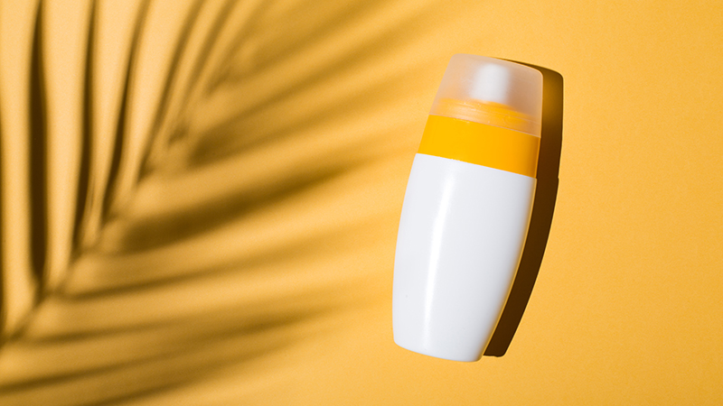 White bottle of sunscreen on a yellow background with a palm leaf shadow over it.