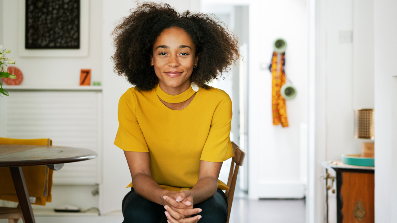 Woman with curly dark brown hair, medium tan skin and gold blouse, smiling into the camera, sitting in a kitchen.