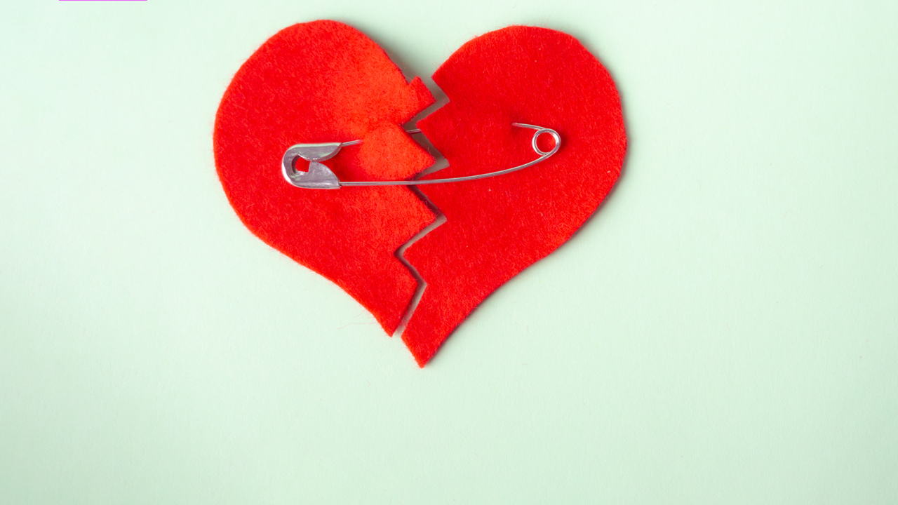 A red felt heart on a light green background. The heart has a zig zag cut down the middle with a safety pin holding it together.