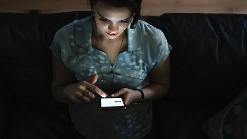 A person looking down at their phone in the dark with light from the phone shining up at their face.