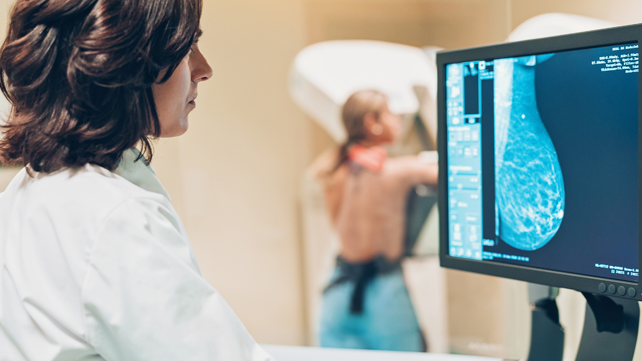 A doctor looks at a computer image of breast tissue while a patient stands at a machine for a mammogram.