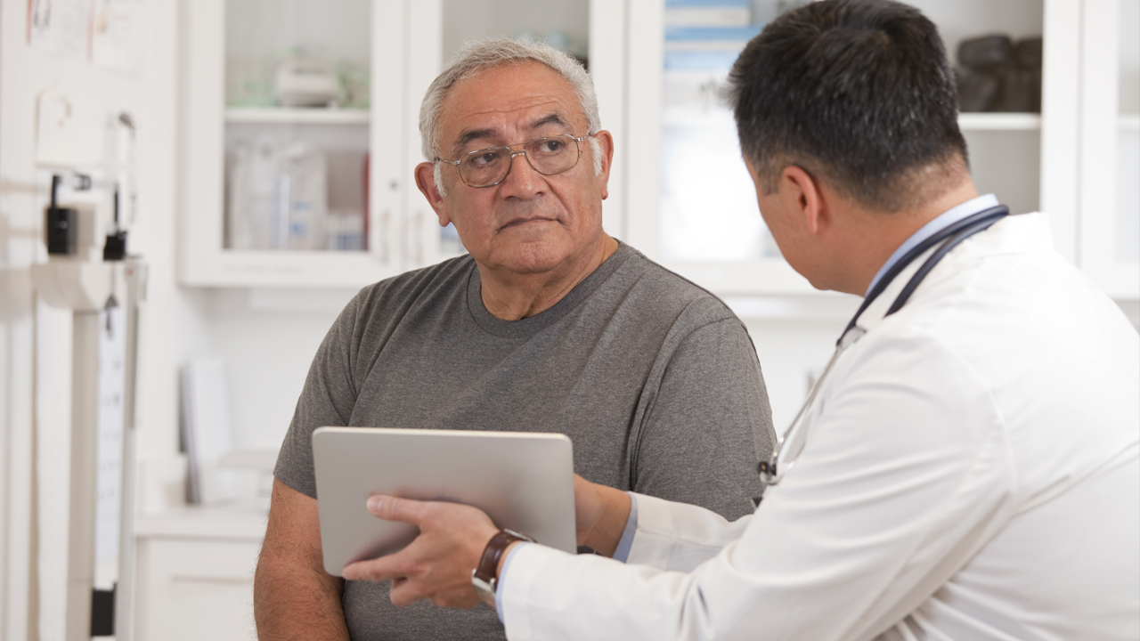 Mature man with pale skin, glasses, white hair, and a gray t-shirt talks with a younger doctor with black hair and a white lab coat, holding a tablet..