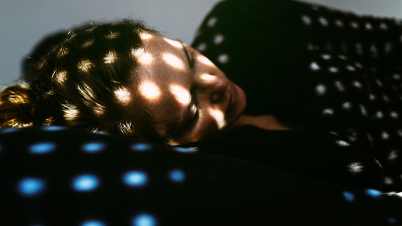 Person asleep with various spots of light casted on their face through gaps in the window blinds.
