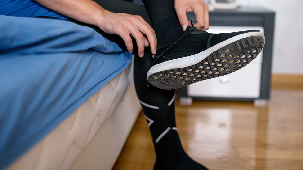 A person wearing black compression socks and taking off his running shoes while sitting on a bed with blue sheets.