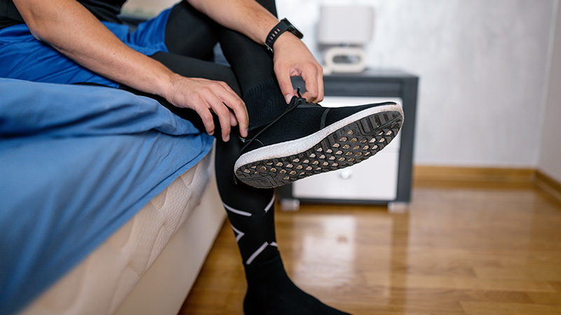 A person wearing black compression socks and taking off his running shoes while sitting on a bed with blue sheets.