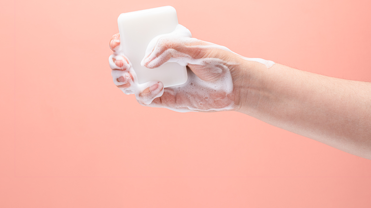 Hand gripping a bar of white soap with white soap suds on a pink background.