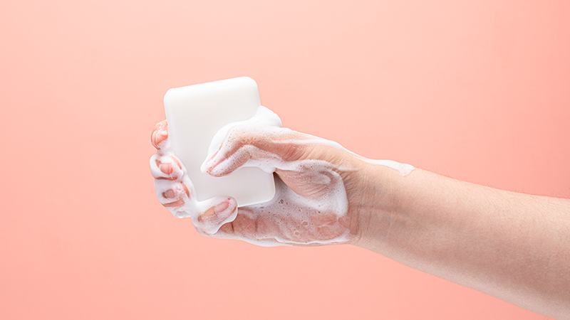 Hand gripping a bar of white soap with white soap suds on a pink background.