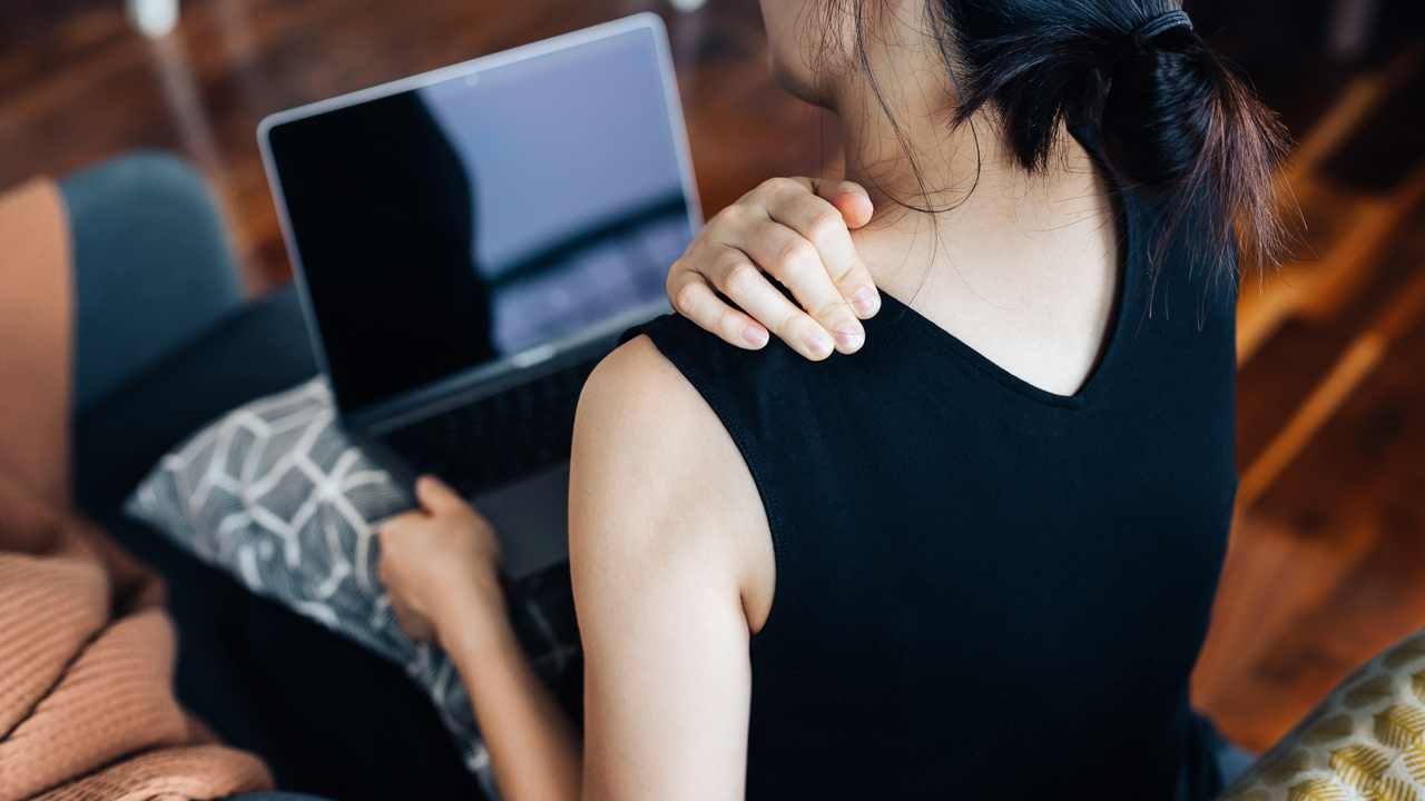 Woman viewed from the back sitting on a couch with a laptop, holding shoulder.