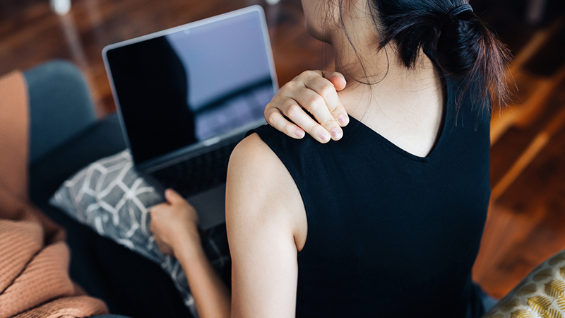 Woman viewed from the back sitting on a couch with a laptop, holding shoulder.