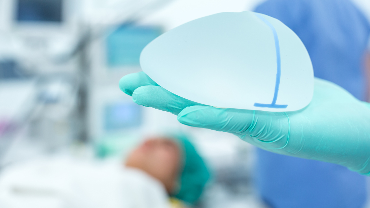 Close up of a hand wearing a blue latex glove holding a breast implant in a surgery room.