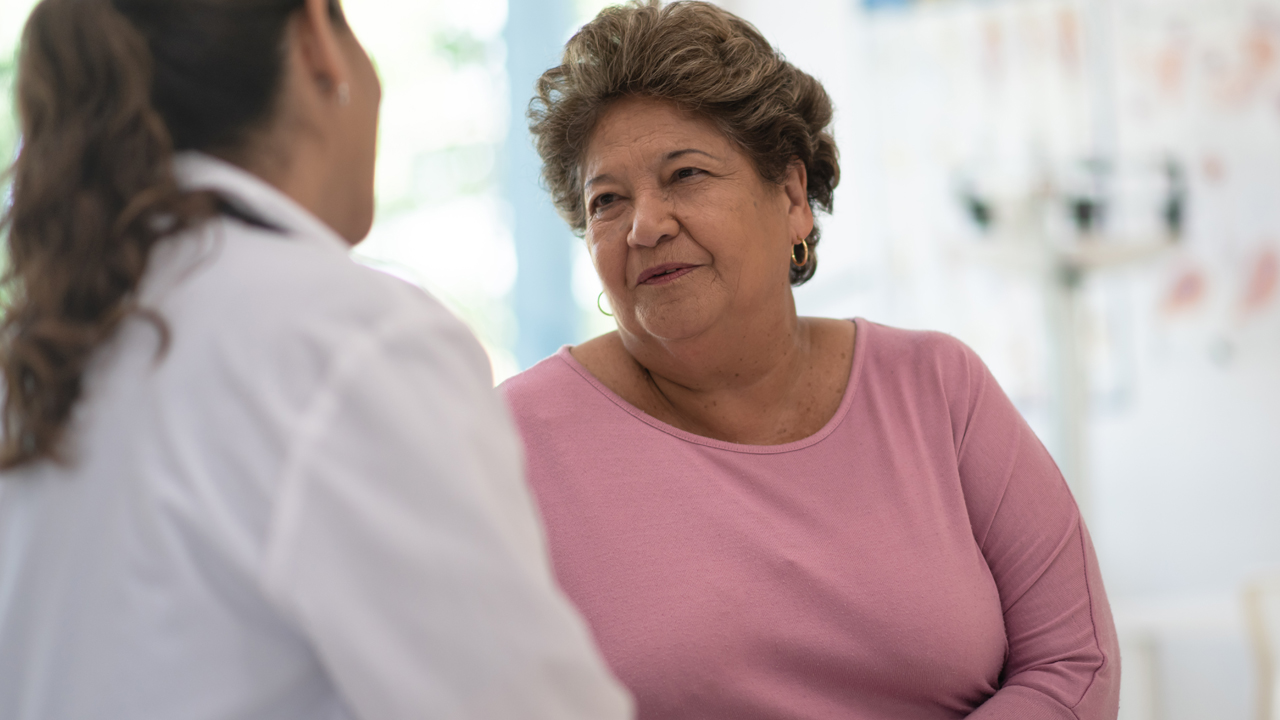 Image taken from behind a physician who is sitting down speaking with a person wearing a long sleeved pink shirt. 