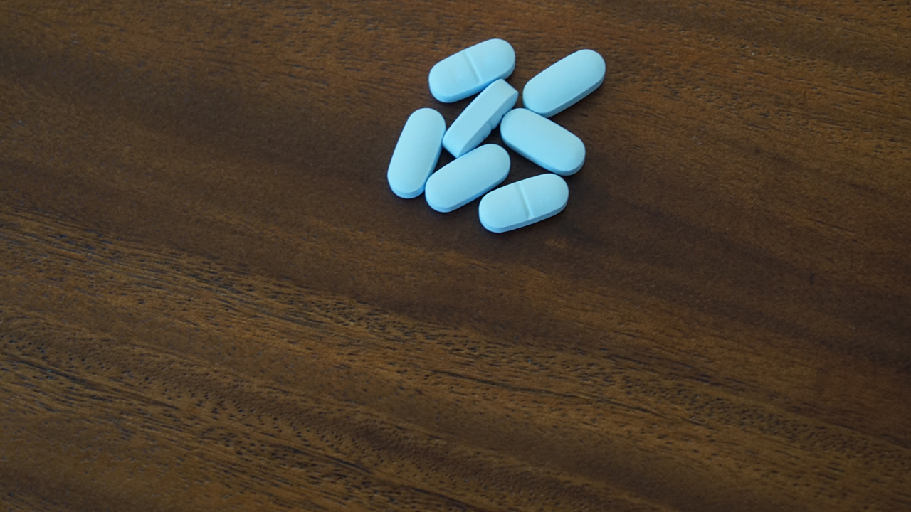 Seven light blue oblong pills in a group on a brown wooden table top.