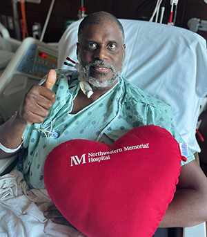 In a hospital bed after surgery, Jerry Dorsey gives a thumbs up while holding a heart-shaped pillow that says Northwestern Memorial Hospital.
