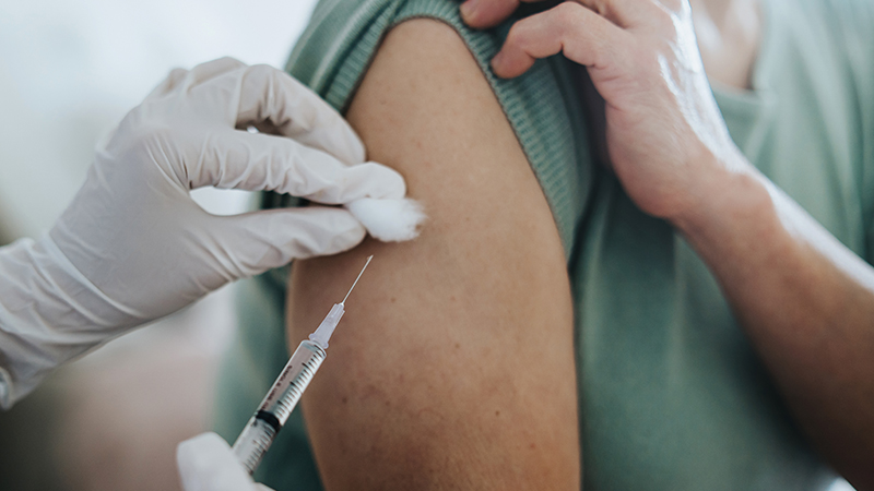 Person holding their sleeve up while another person wearing a white rubber glove holds a syringe and needle in one hand and a cotton ball in another. 