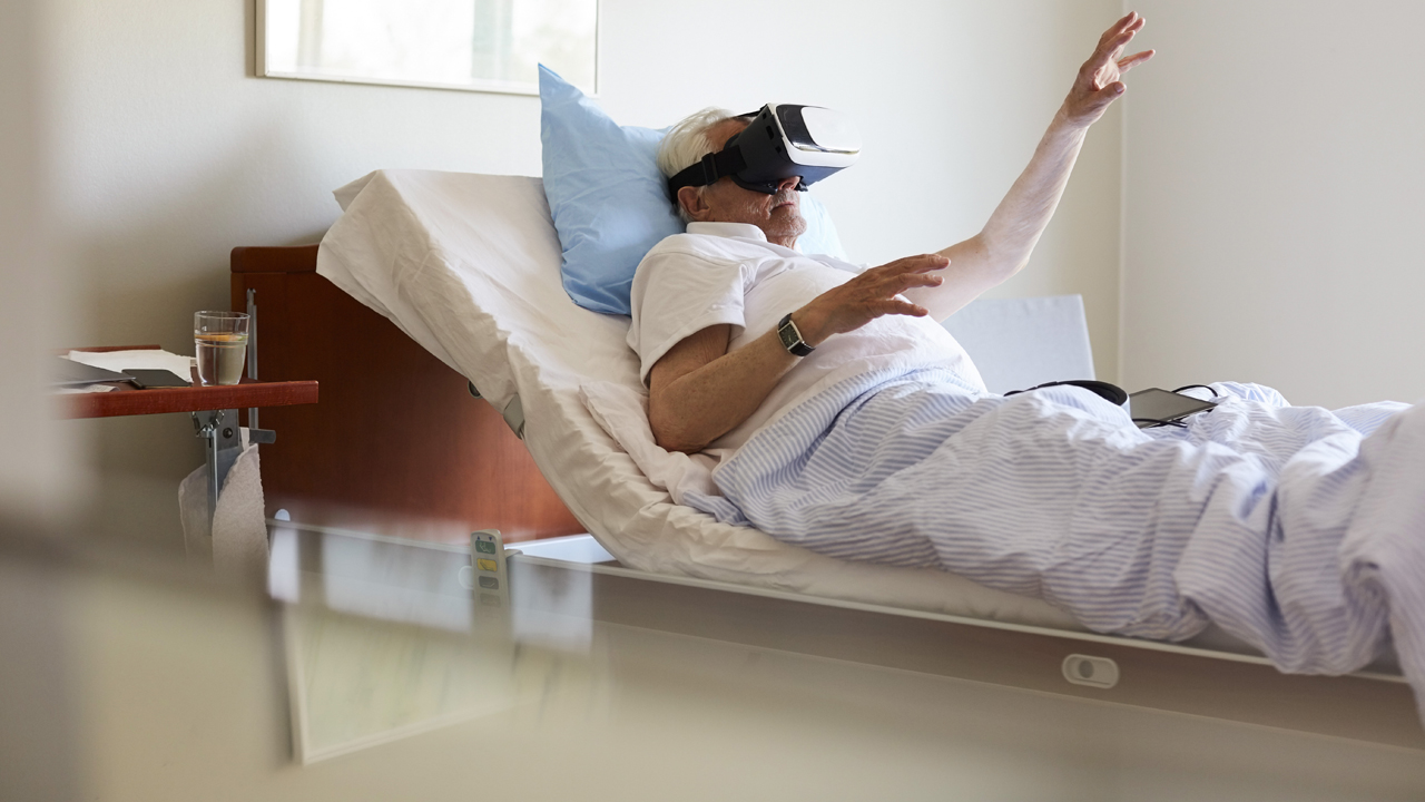 Senior man gesturing while using VR glasses on bed in hospital.