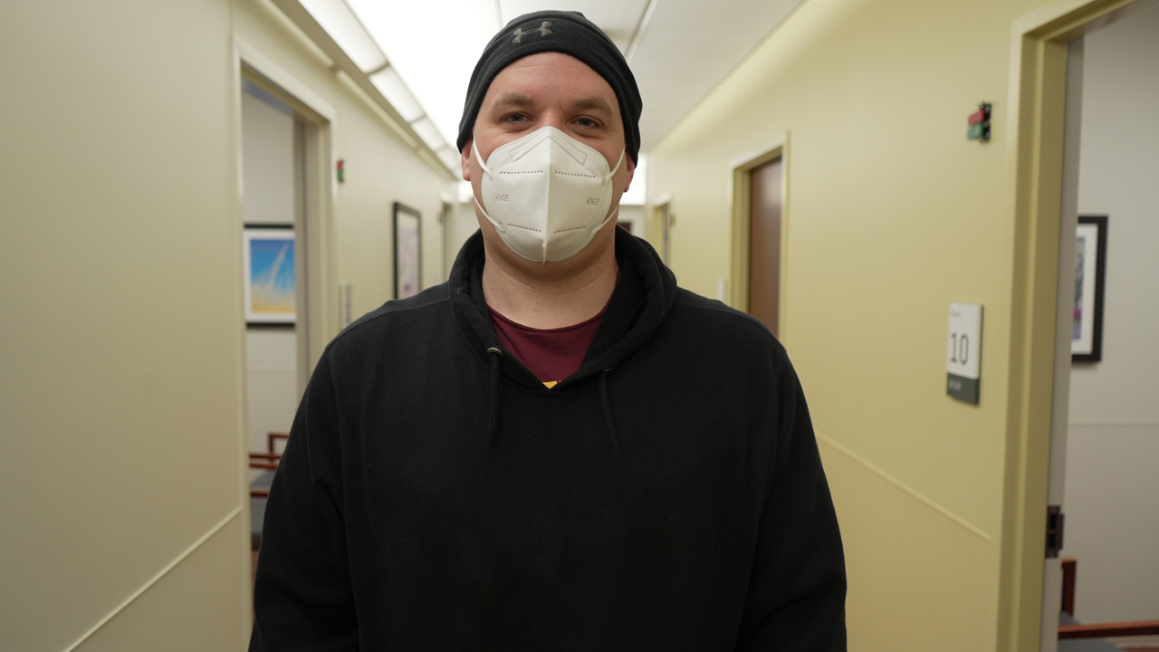 A person in a surgical mask wearing a black hoodie stands in a hospital hallway. 