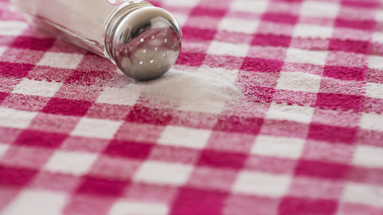 A salt shaker turned on its side with salt poured out on a red checked tablecloth.