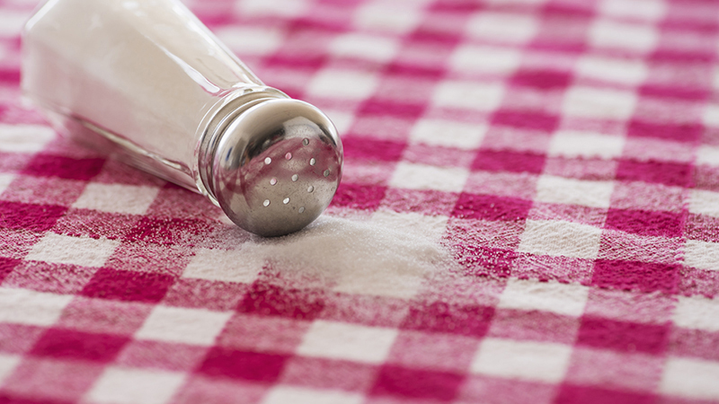 A salt shaker turned on its side with salt poured out on a red checked tablecloth.