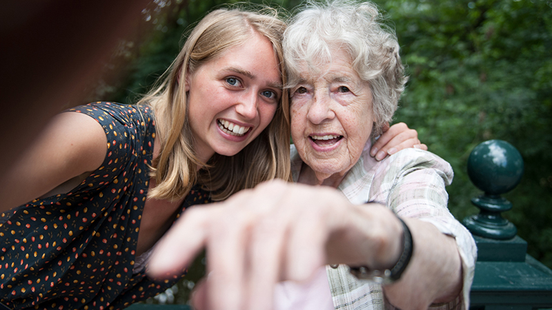 Young woman with light skin and long blond hair takes a selfie with an elderly woman with pale skin and white curly hair, looking into the camera smiling.