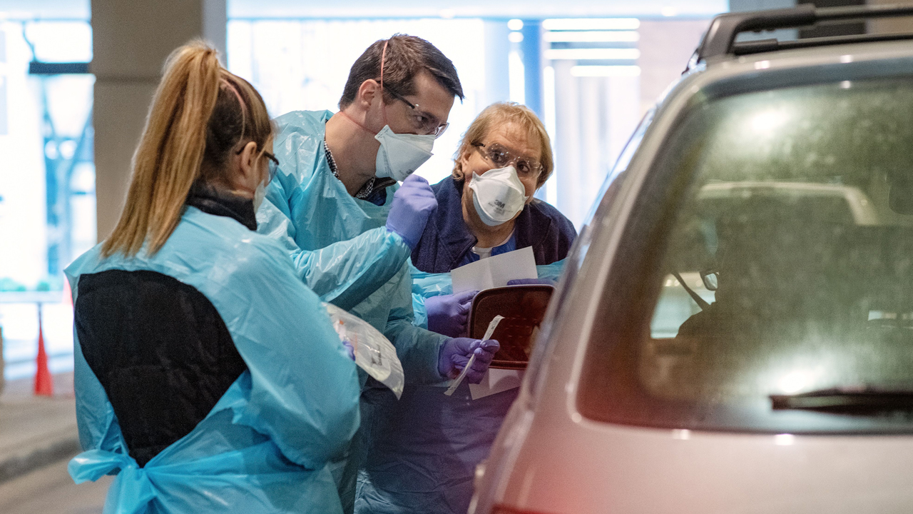 Three clinicians conduct a COVID-19 test outside a gold vehicle. 