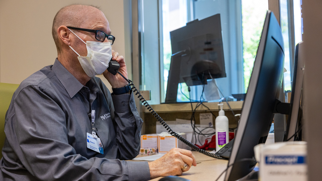 Edward Hogan using a landline sitting at a desk in front of a computer. He is balding and wears glasses, a white surgical mask, and a dark gray shirt with a Northwestern Medicine logo and badge.