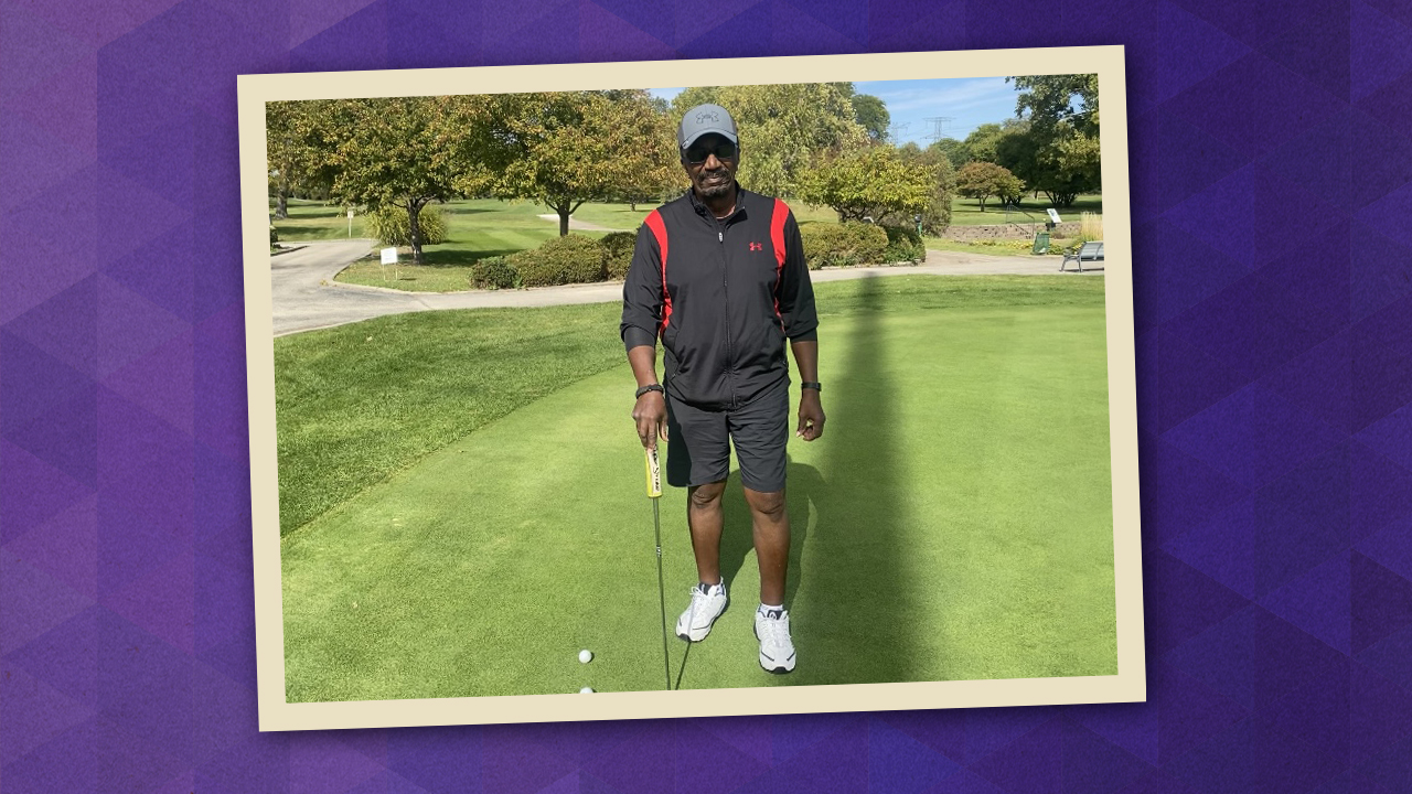 Sidney Strong pictured on a putting green holding a golf club and wearing black shorts, black and red jacket, sunglasses, and gray ball cap.