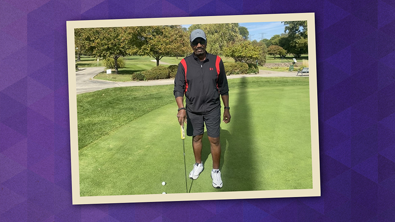Sidney Strong pictured on a putting green holding a golf club and wearing black shorts, black and red jacket, sunglasses, and gray ball cap.