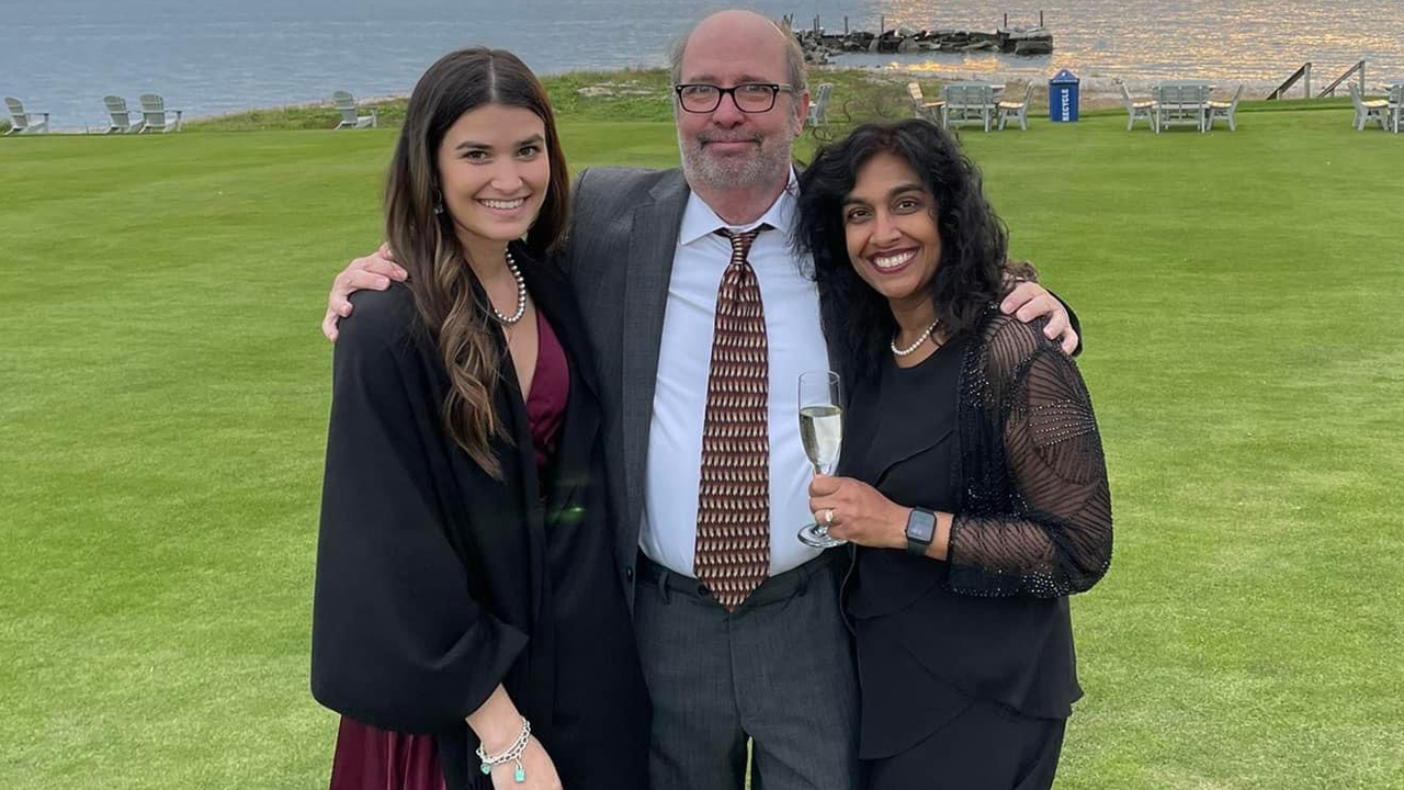 Mark Dybas wears glasses, shirt, a tie and blazer. He is pictured center with his wife and daughter on either side of him. They are all on a green lawn with a lake and sunset behind them.