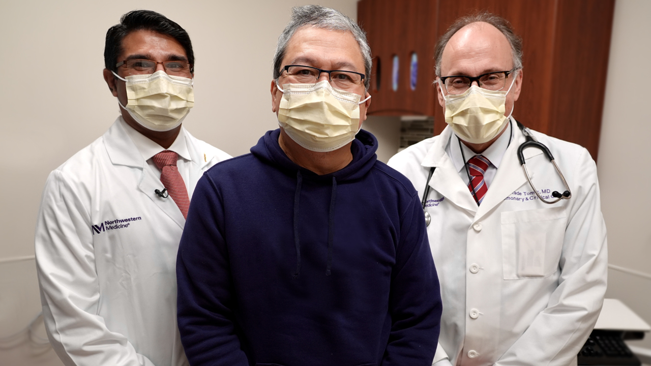 Patricio Collera wearing a dark blue sweatshirt and yellow surgical mask, with his lung-liver transplant surgeons on either side, who wear white lab coats and yellow surgical masks.