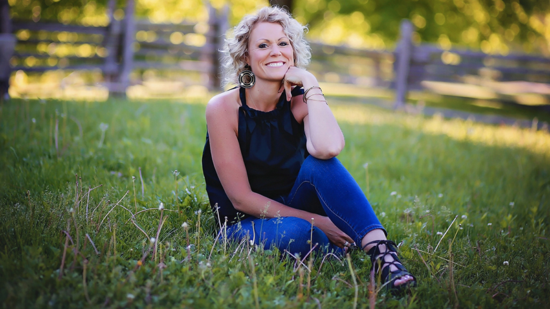 Centered in the photo, Sherri Kalmer smiles while sitting on a lawn. She is blonde and white, and she wears a black sleeveless top and blue jeans. 