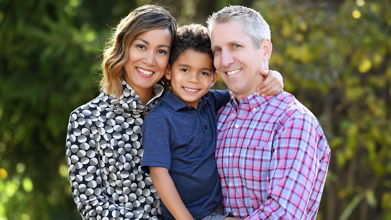 Family portrait taken outside in front of foliage of mother, son and father. Mother is on the left, wearing a black and white dress. Son is in the middle being held by dad. Son is grade school age and wearing a navy polo. Father is on the left wearing a plaid button-down shirt. 