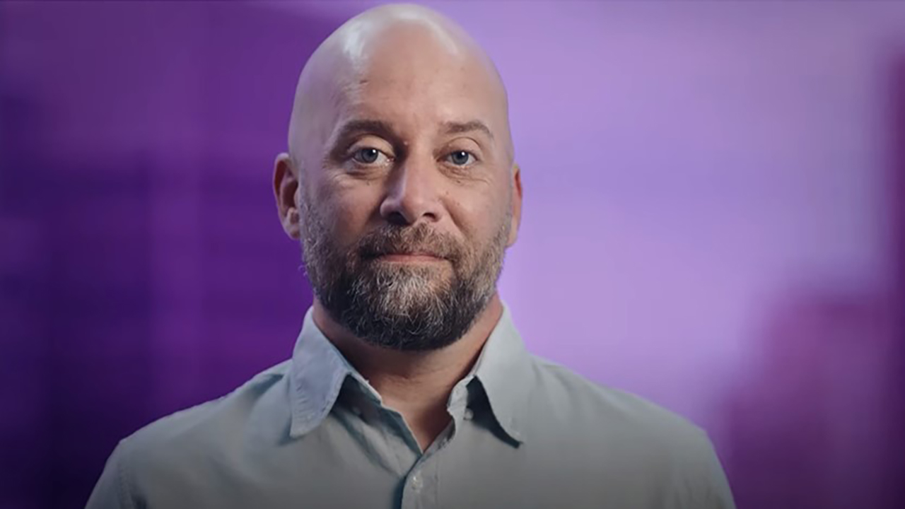Phil Rister poses in front of a vibrant purple background. He is visible from the shoulders up. 