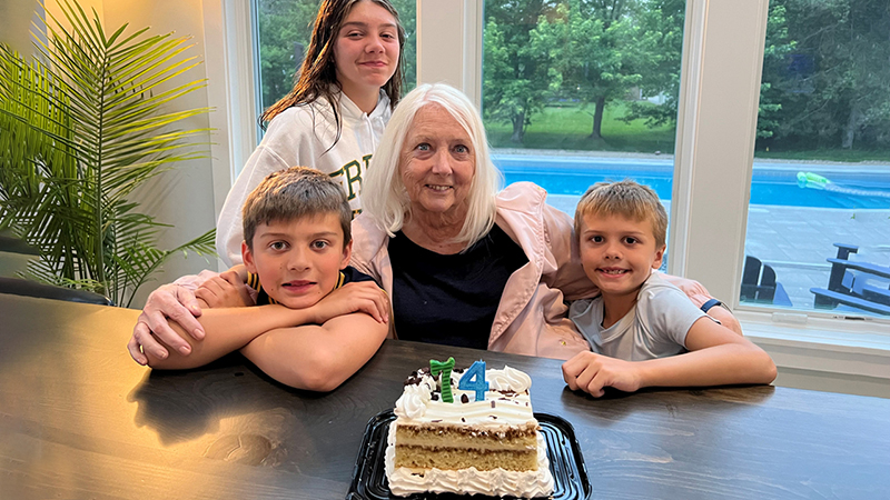 Barbara Le Breton, with shoulder length white hair and and wearing a black blouse and pink jacket, has her arms around two boys at a table. A girl smiles in the background. There is a birthday cake on the table in front of them with candles shaped like "74."