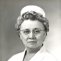 Black and white headshot of Bertha Knapp, RN. She is a white woman wearing a white nurses' cap, a white shirt with a pin on it and has curly hair and glasses. 